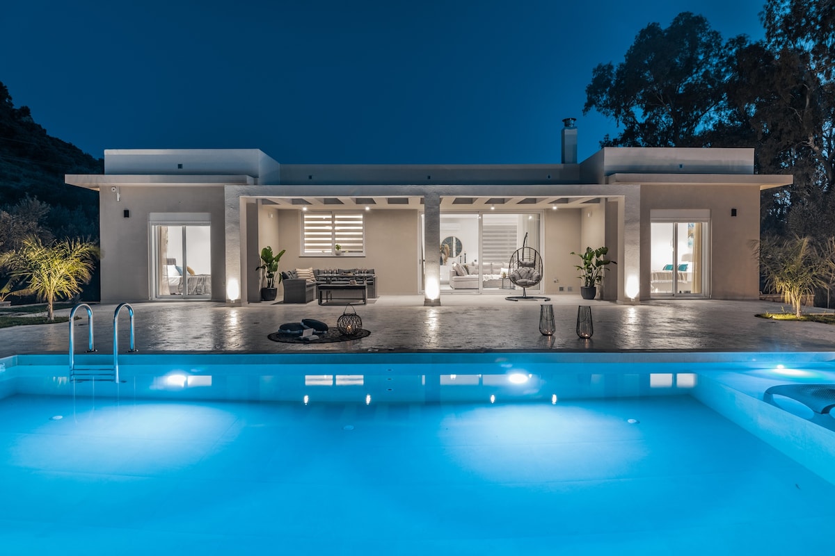 The villa is showcased at night, illuminated softly by strategic lighting. A serene swimming pool reflects the blue hues, while the spacious terrace is lined with modern seating areas. Large glass doors provide a view into the inviting interior spaces, enhancing the overall tranquil ambiance.