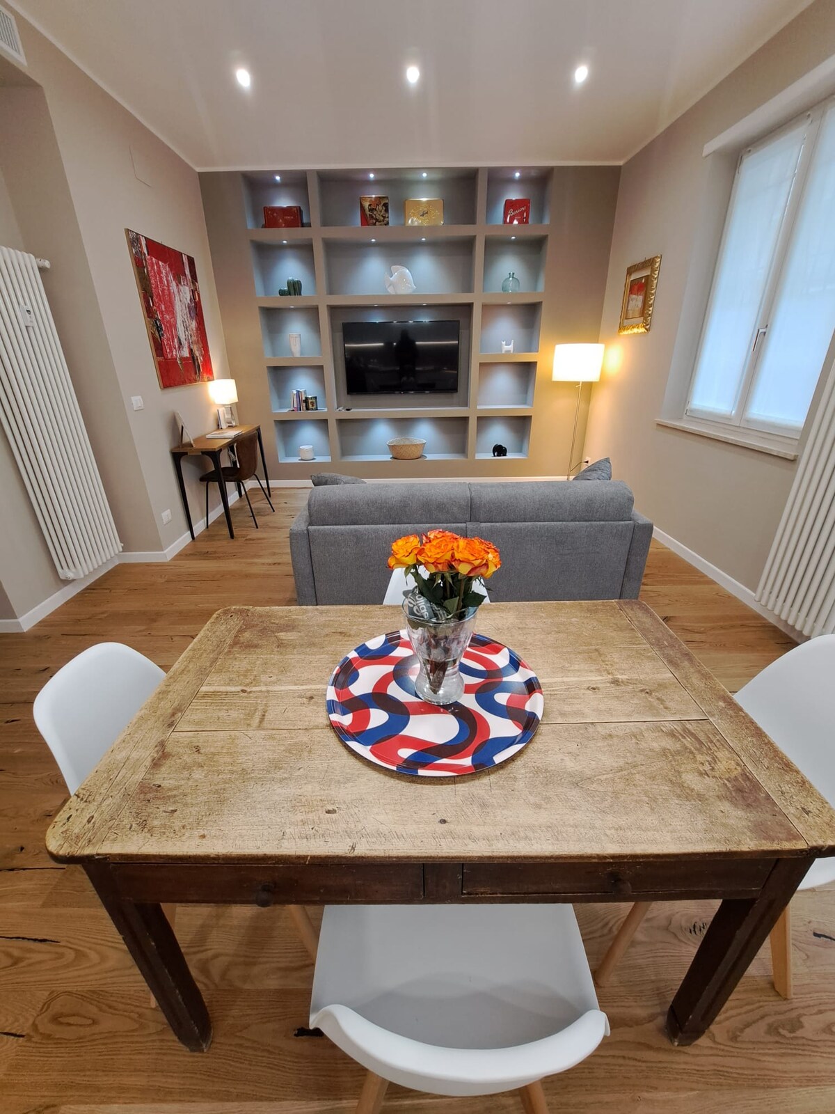 A cozy living space is highlighted by warm wooden flooring and a rustic dining table with a decorative tray and fresh flowers. A grey sofa is positioned near a built-in shelving unit, which features lighting and a flat-screen TV. Soft lamps create a welcoming ambiance.