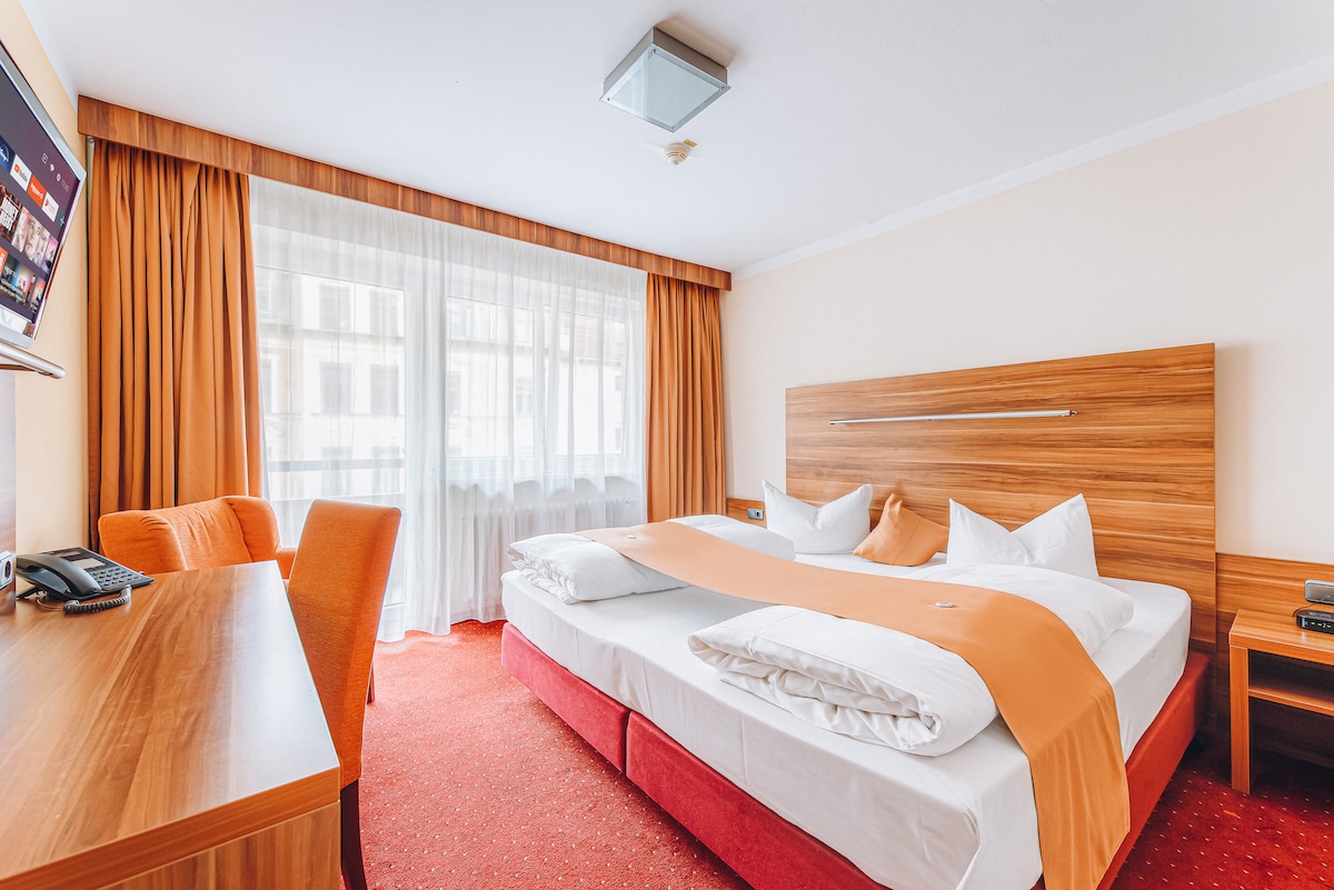 A spacious room features a large bed with crisp white linens and a bold orange accent. Natural light floods the area through large windows draped with light curtains. A cozy seating area is highlighted by an orange armchair, and a wooden desk offers a workspace.