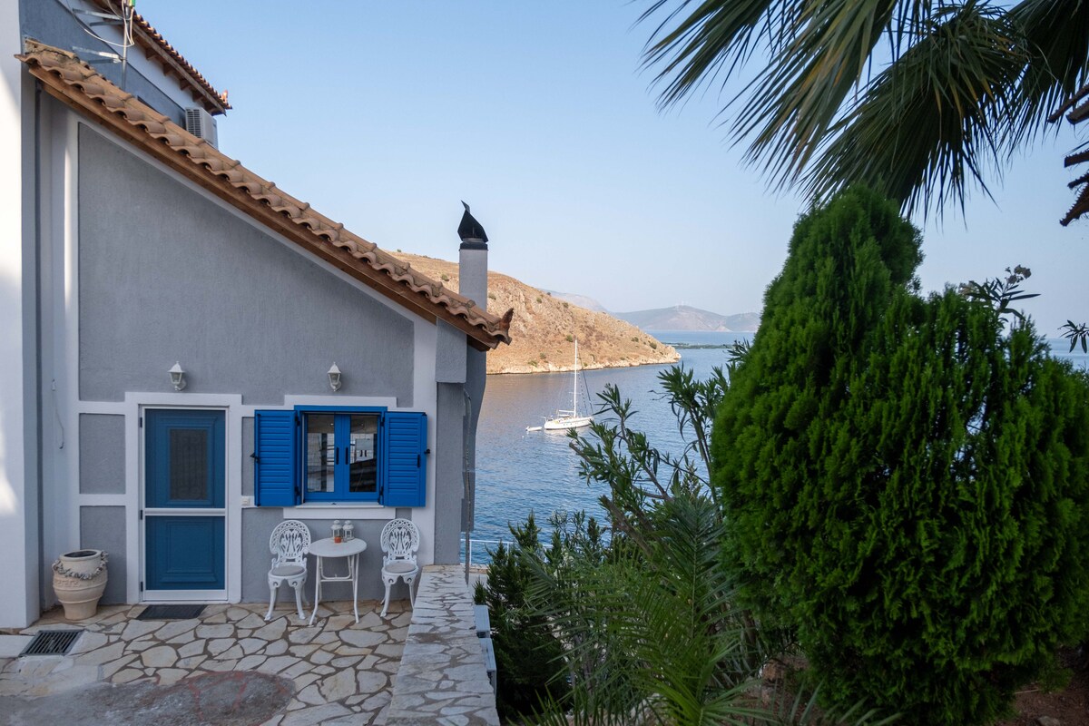 A charming exterior view features a two-story residence with blue shutters. A small seating area with two white chairs is positioned outside, inviting relaxation while overlooking the tranquil sea and distant hills. Lush greenery and palm trees frame the scene, enhancing the natural beauty.