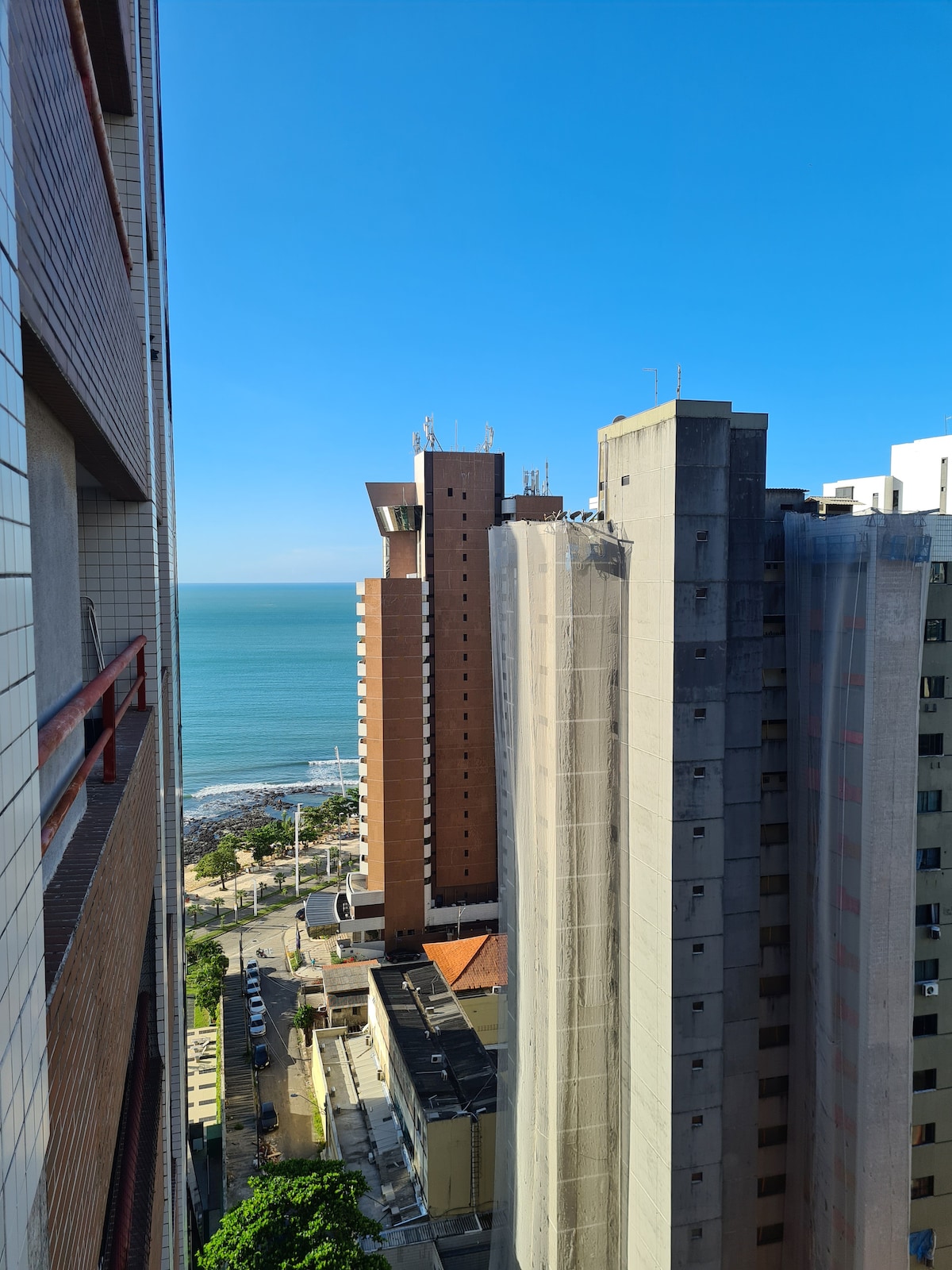 A view from a balcony showcases the tranquil blue sea and sandy beach, positioned below tall buildings. Clear skies complement the coastal scenery, with a glimpse of the bustling street below. The buildings nearby exhibit a mix of textures and materials, creating a vibrant urban atmosphere.
