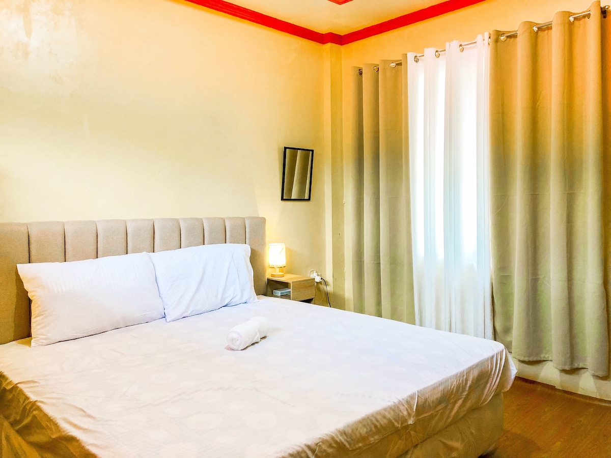 A spacious bedroom features a comfortable bed with a white sheet and a rolled towel placed on top. Soft curtains frame a pair of windows allowing natural light to brighten the room. A small bedside table is positioned next to the bed, holding a lamp and a mirror.