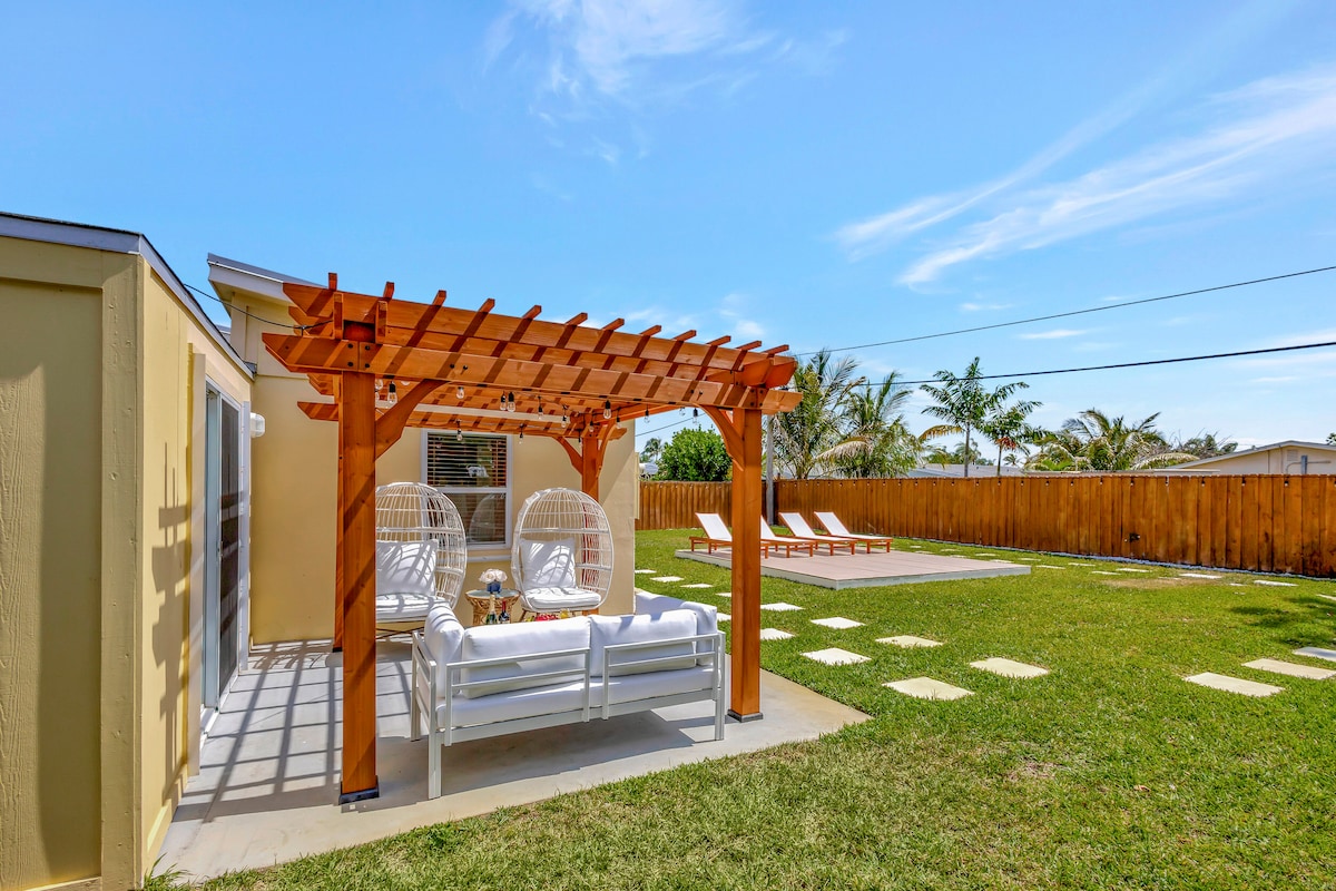 An outdoor space features a wooden pergola with hanging chairs and a comfortable seating area. A manicured lawn is visible, accompanied by lounge chairs positioned for relaxation. The clear blue sky and spacious layout provide an inviting atmosphere for outdoor gatherings.