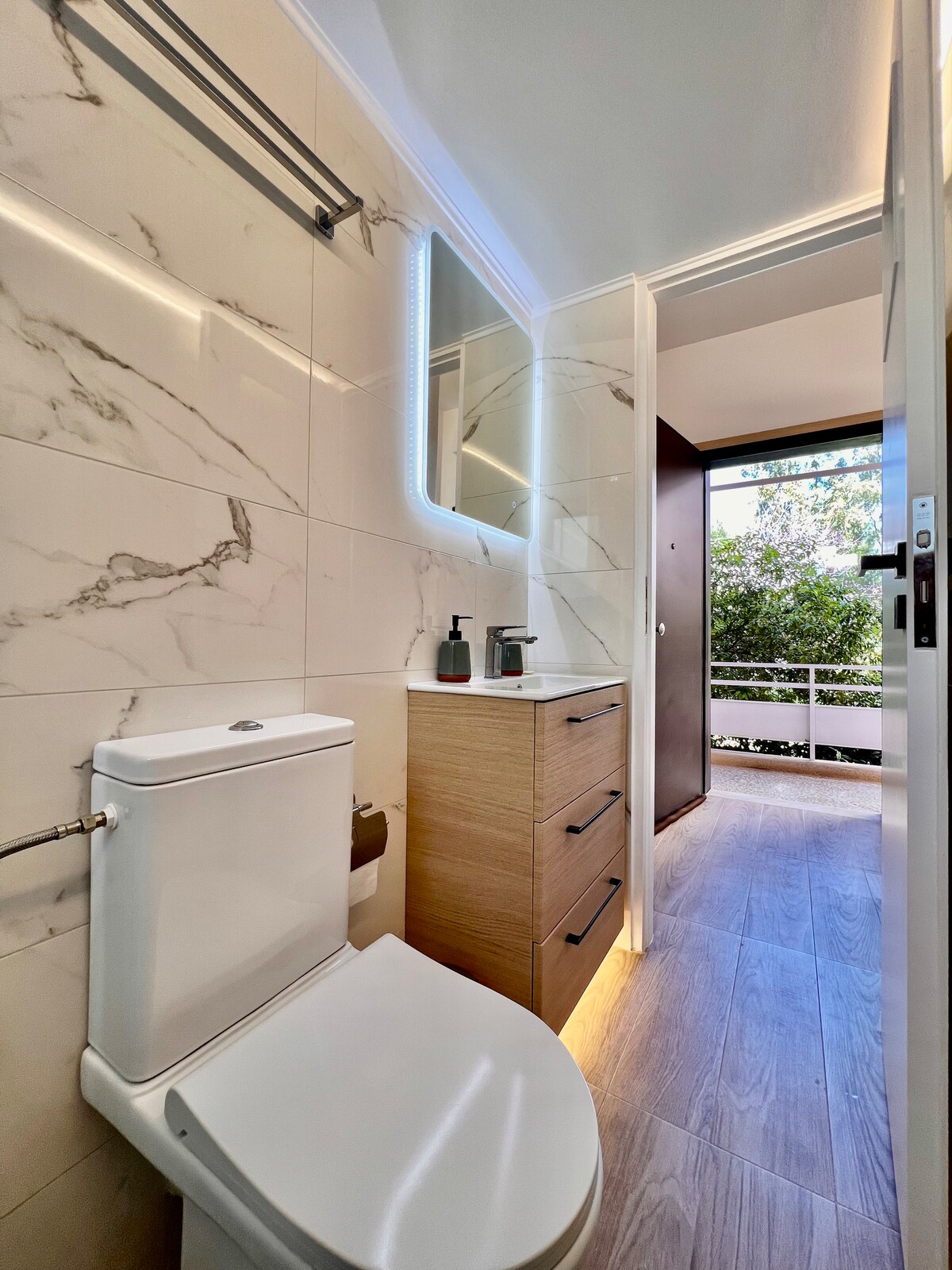 A modern bathroom features sleek marble walls and a compact vanity with several drawers. Soft lighting is provided by a well-placed LED mirror, and a view of greenery is visible through the open door, enhancing the overall ambiance.