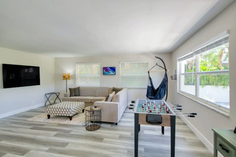 A spacious living area features a comfortable sectional sofa and a flat-screen TV mounted on the wall. Natural light streams through large windows, illuminating a foosball table and a decorative coffee table that anchors the space.