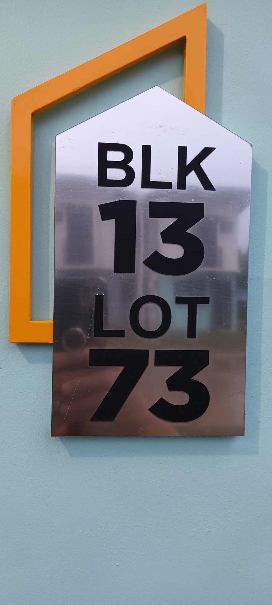 A metallic sign features clear, bold numbers indicating 'BLK 13 LOT 73,' attached to a light blue wall. An orange geometric shape accentuates the sign, providing a contemporary touch. The overall design ensures visibility, guiding guests to their destination.