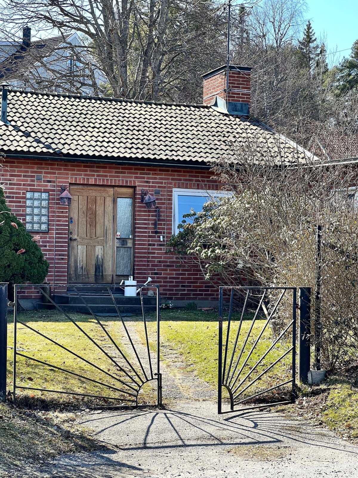 A brick villa is framed by a black wrought-iron gate leading to a grassy pathway. The entrance features a wooden door with glass panels and two lanterns on either side. Lush greenery surrounds the property, enhancing the inviting exterior.