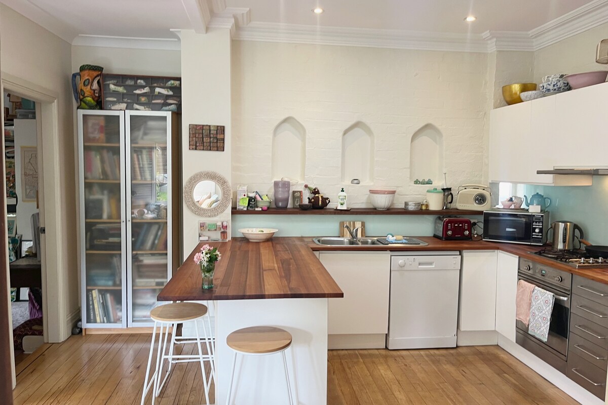 A bright kitchen area features a wooden countertop with two bar stools positioned at one side. Modern appliances, including a dishwasher and microwave, are neatly arranged. Light streams in from a nearby window, highlighting the vintage decor on the shelves and the white brick walls.