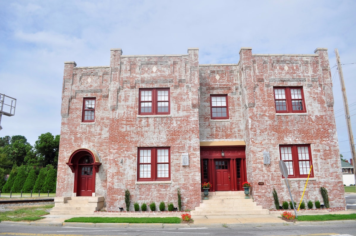 A historic brick building is showcased, featuring a symmetrical facade with large windows framed in red. The entrance is highlighted by a rounded arch and steps leading to the front door. Lush greenery aligns the walkway, contributing to the welcoming appearance.