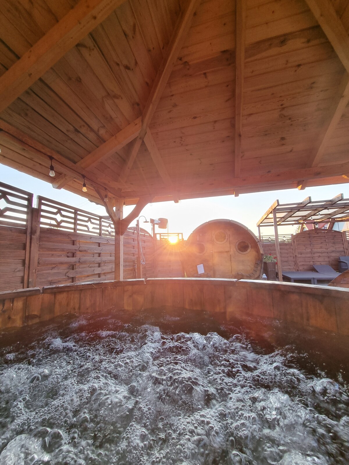 A wooden gazebo shelters a hot tub, with bubbles creating a frothy surface. Light streams in from the sunset, casting a warm glow. Wooden walls surround the area, providing privacy, while soft lighting enhances the tranquil ambiance.