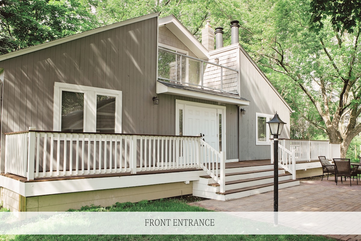 The front entrance of the home is framed by a spacious deck, featuring a light post and seating area. Large windows provide natural light, while the neutral-colored siding complements the surrounding greenery.