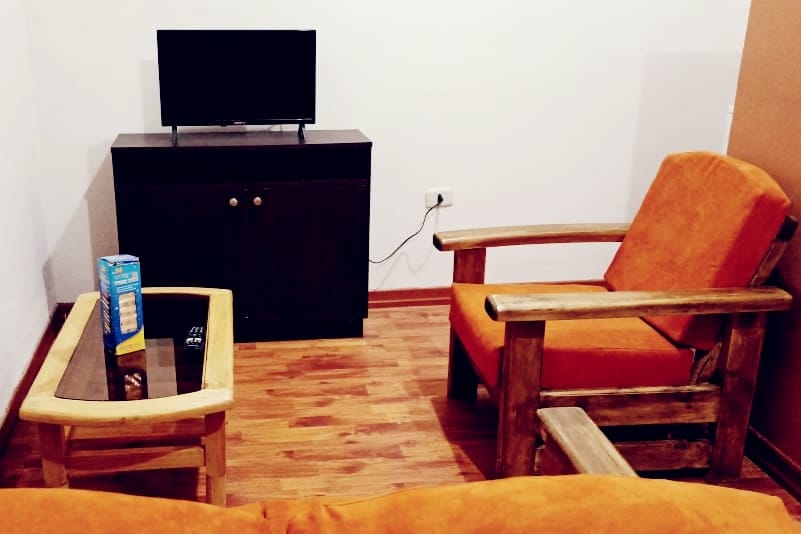 A cozy living area features a wooden coffee table, complemented by an orange armchair and a matching sofa. A TV is placed on a dark wooden cabinet against the wall, with wooden flooring accentuating the warm tones.