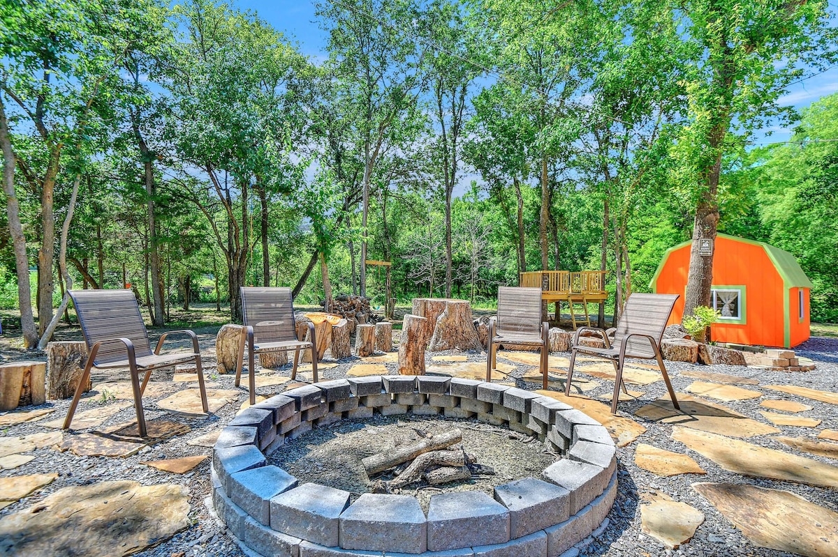 A circular fire pit area is surrounded by stone seating and natural stone pathways, integrated into a shaded outdoor space. Lush greenery and trees provide a serene backdrop, with a vibrant orange structure visible in the distance, enhancing the spacious and inviting atmosphere.