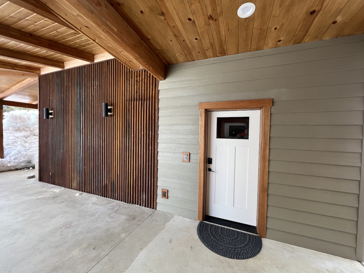 The exterior entrance features a modern door framed with warm wooden accents. The walls showcase a combination of textured wood and smooth siding. The covered porch is complemented by two wall sconces providing soft lighting, enhancing the welcoming appearance.
