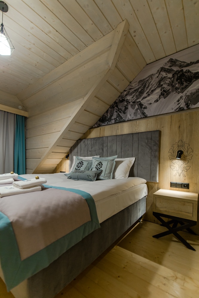A serene bedroom features a spacious bed adorned with decorative pillows. Soft colors are present in the textiles, while wooden paneling and sloped ceilings create an inviting atmosphere. A nightstand with a subtle lamp is positioned beside the bed, complementing the overall design.