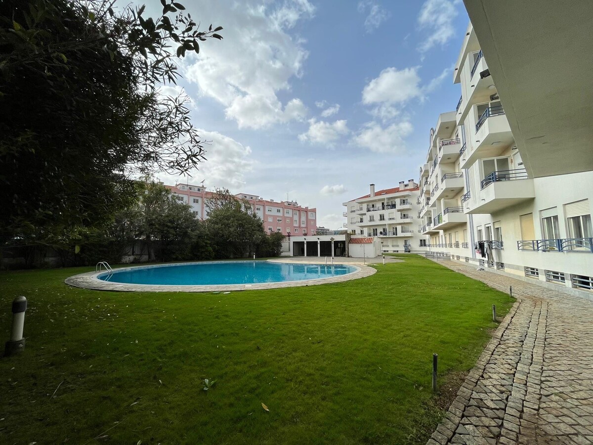 A spacious communal area features a large swimming pool surrounded by green lawn. White buildings with balconies are visible in the background, offering a serene setting. The area is illuminated by natural light, highlighting the inviting pool space.