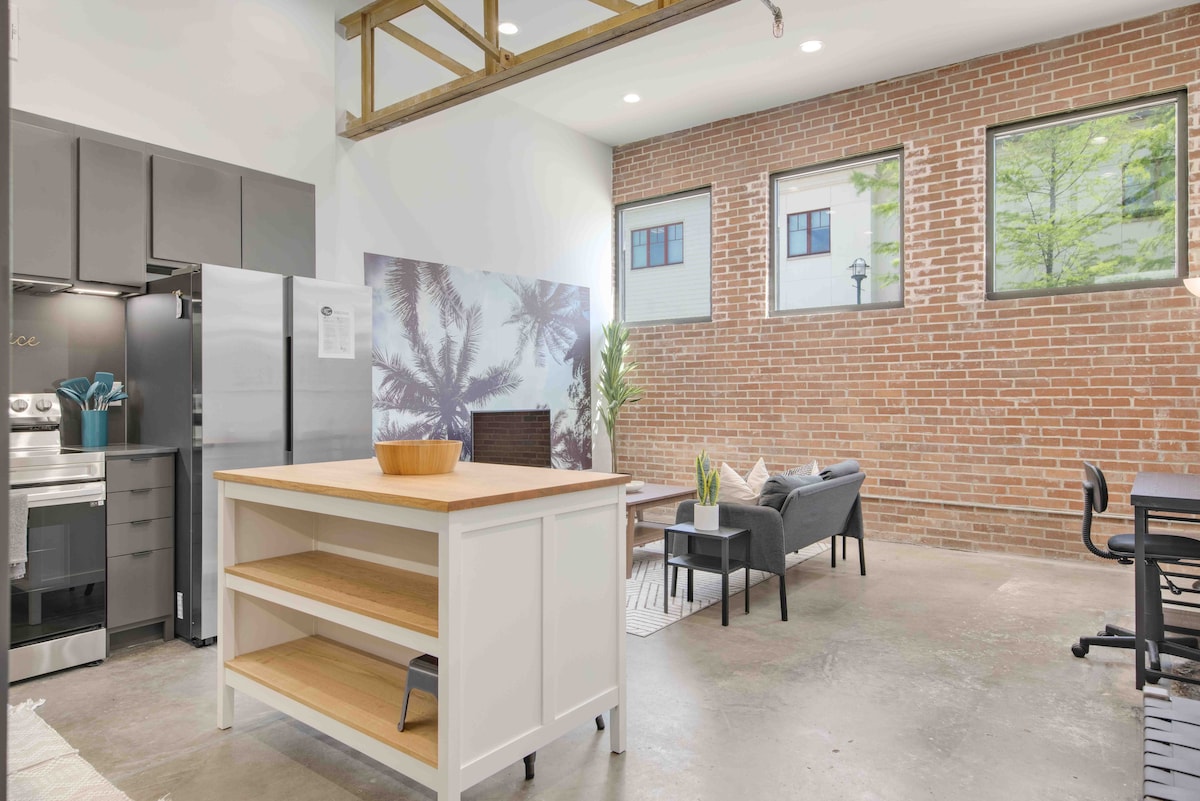 The modern kitchen features a central island with open shelving, ideal for meal prep. A spacious living area is visible, adorned with a comfortable seating arrangement and large windows providing natural light. The brick wall adds a touch of industrial charm to the space.