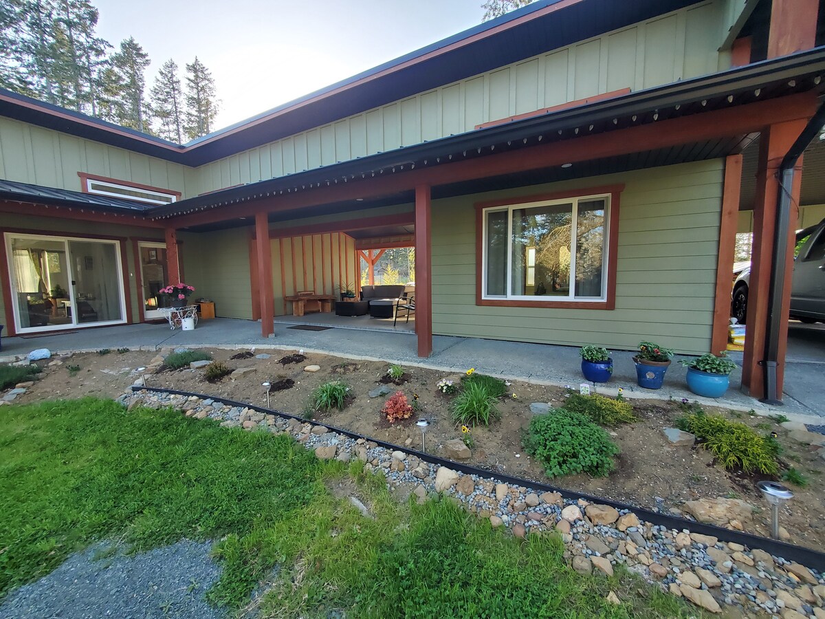 The exterior of the property features a covered porch area with seating and a well-maintained garden bed filled with various plants. Large windows provide views of the inviting outdoor space, while the surrounding landscape includes greenery and trees, creating a serene environment.