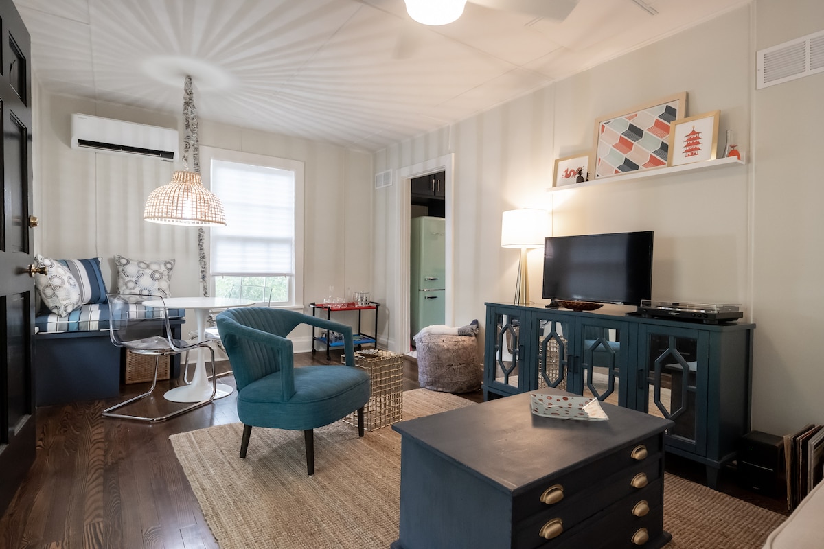 The living area features a comfortable blue chair and a stylish coffee table, with a flat-screen TV positioned atop a decorative cabinet. A bright light fixture hangs above a dining table, complemented by a cozy seating nook near the window. Warm wood flooring adds to the inviting ambiance.