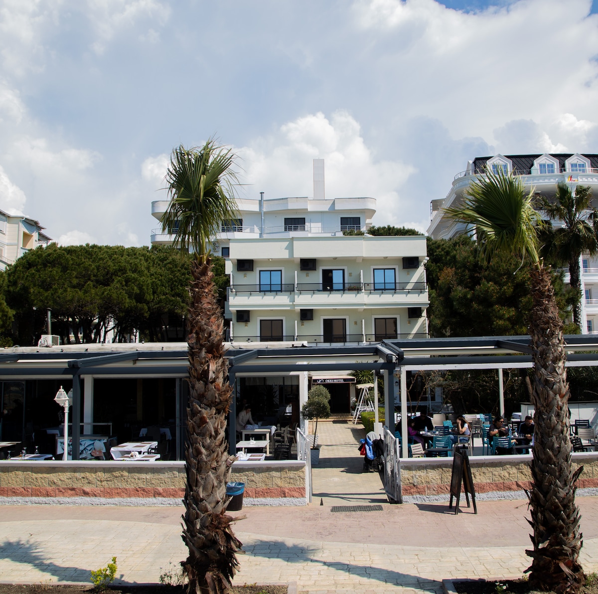 The hotel exterior is framed by tall palm trees, with a modern structure featuring multiple balconies. A spacious outdoor seating area is visible in front, offering tables and chairs under a covered patio. A clear sky with scattered clouds is overhead.