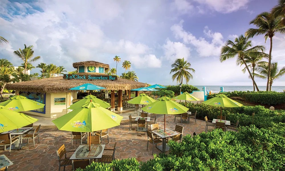 A casual bar is showcased with bright green umbrellas providing shade over wooden tables and chairs. Lush greenery runs along the stone walkway that leads to the beachside. The ocean is visible in the background, with palm trees gently swaying under a partly cloudy sky.