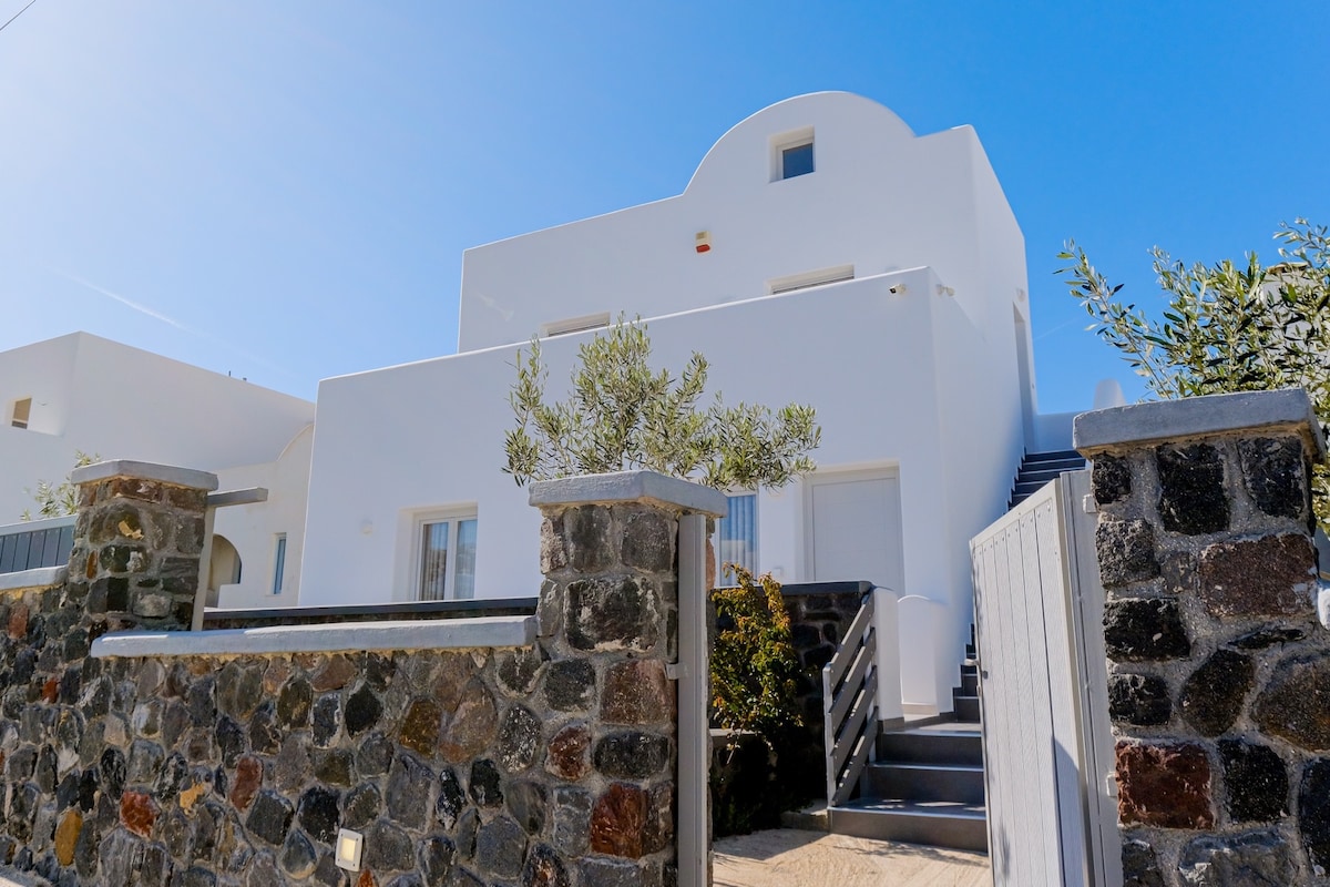 The exterior of Ageri Suite is presented, showcasing a sleek, whitewashed building characteristic of Santorini architecture. The entrance is framed by a stone wall, with stairs leading to the second floor, surrounded by greenery under a clear blue sky.