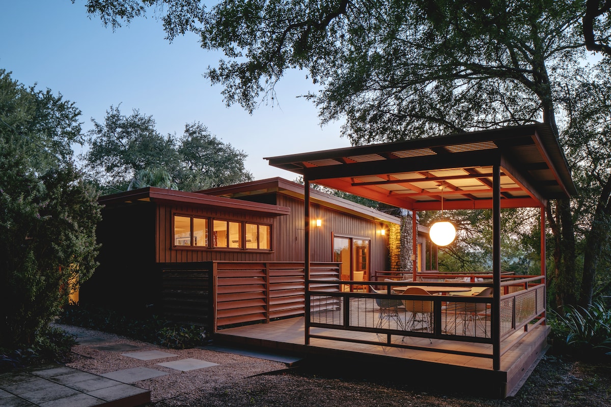 A modern exterior view of the home showcases a deck featuring a dining area under a canopy. Large glass windows illuminate the interior, while surrounding trees provide a natural backdrop. The warm evening light creates a welcoming ambiance in this secluded setting.