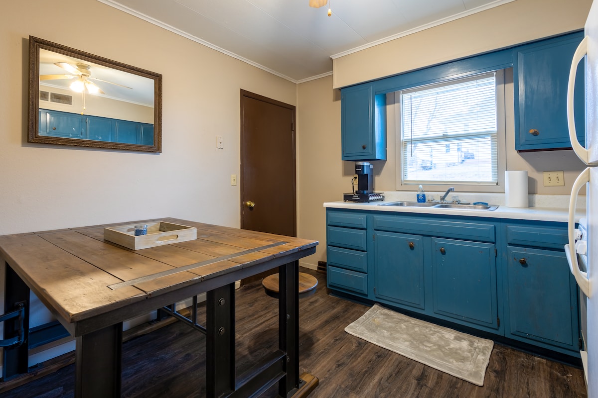 A well-equipped kitchen features blue cabinets and a window that allows natural light. A wooden dining table with a decorative tray is positioned in the center, while a countertop displays essential appliances, including a coffee maker. A soft rug lies on the floor near the appliances.