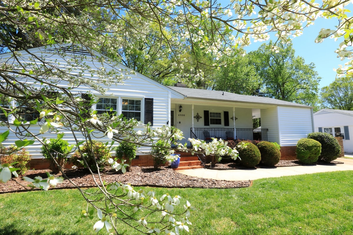 A charming single-story home is presented, featuring a white exterior and a covered front porch. Lush green grass surrounds the property, complemented by flowering trees and neatly trimmed bushes. A pathway leads up to the entrance, offering a welcoming approach.