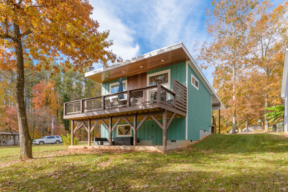 A modern lake house is set against a backdrop of autumn foliage, featuring a spacious deck with seating. The light blue-green exterior complements the natural surroundings, while large windows reflect the inviting atmosphere of the home.