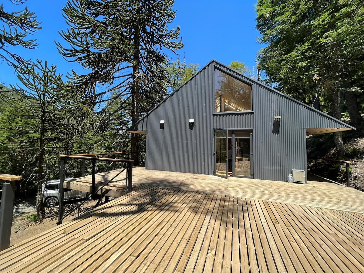A modern cabin is presented with a sloped roof, featuring large windows that invite natural light. A spacious wooden deck surrounds the entrance, providing a seamless connection to the natural environment. Tall trees are visible in the background, contributing to the serene setting.
