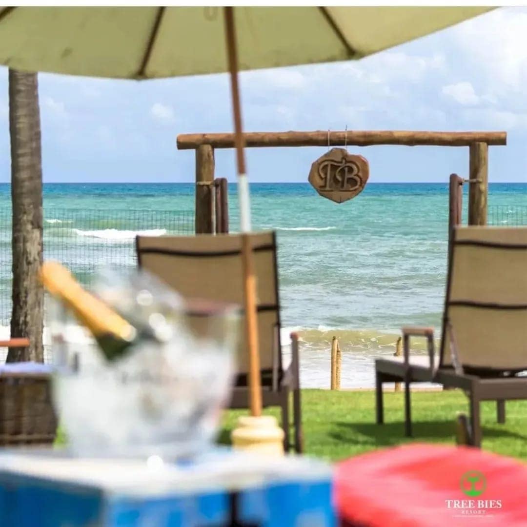 A serene beachfront is presented, with inviting loungers arranged under an umbrella. A wooden sign with the initials 'TB' is visible near the shore, overlooking the calm, blue ocean waters under a partly cloudy sky.