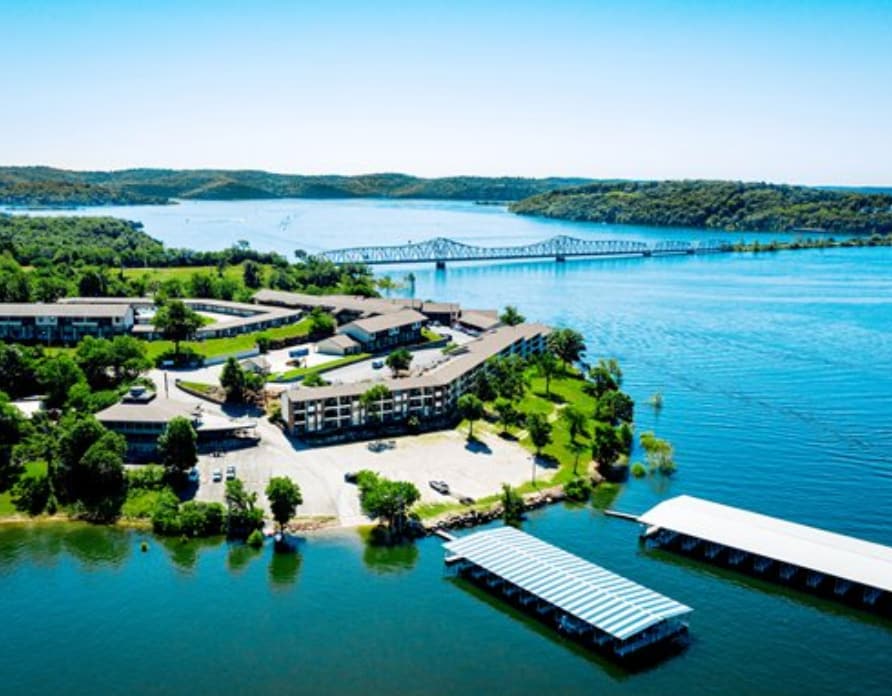 Aerial view of the Kimberling Inn Resort, positioned along Table Rock Lake. Lush green grounds blend into the water, which reflects the clear blue sky. Boat slips and a large parking area are visible, alongside a bridge in the distance, creating a scenic lakeside setting.