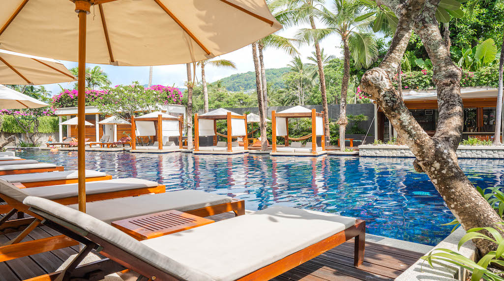 A serene outdoor pool area features lined sun loungers with beige cushions under large umbrellas. Lush greenery surrounds the space, with palm trees and flowering plants adding natural beauty. The calm blue water reflects the clear sky and provides a relaxing atmosphere.