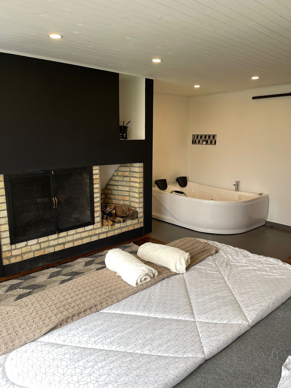 A cozy bedroom features a spacious bed covered with a neutral quilt and two rolled towels placed neatly on top. A modern bathtub is positioned nearby, complemented by a stylish fireplace made of brick, creating a tranquil setting.