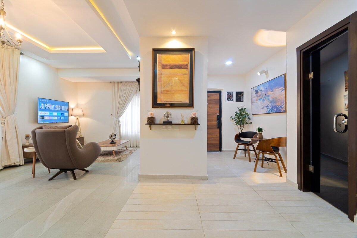 An elegant entrance area features light-colored tile flooring and soft lighting. A comfortable armchair is positioned beside a small wooden table. Artwork adorns the walls, and a large TV is displayed in the adjoining living space. Sheer curtains frame a window allowing natural light to fill the area.