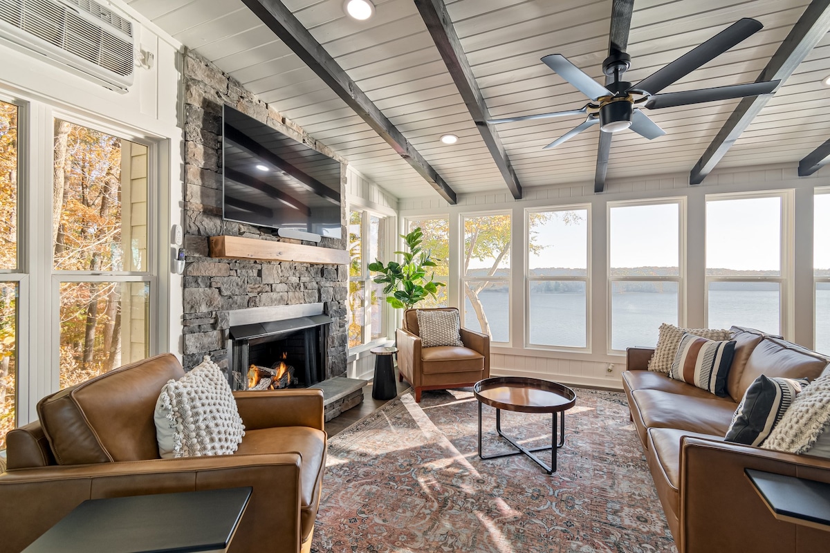 A bright living area features large windows showcasing expansive lake views. Two leather sofas are positioned around a coffee table, with a cozy fireplace and a mounted television on a stone wall. A potted plant adds a touch of greenery to the space.