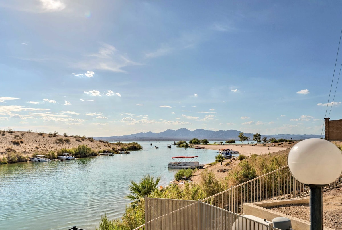 A serene view of the waterway is presented, showcasing a gentle curve lined with sandy shores and sparse vegetation. Small boats can be seen on the water, and distant mountains add depth to the horizon. The scene is illuminated by natural light under a clear blue sky.