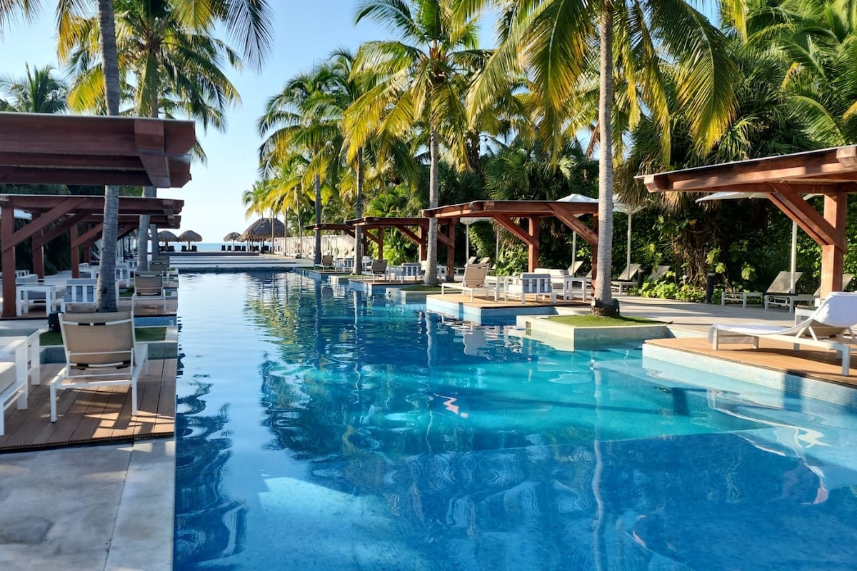 A tranquil pool area is surrounded by tall palm trees, with wooden cabanas providing shaded seating. The clear blue water reflects the sunlight, creating a serene atmosphere. Lounge chairs line the sides, inviting relaxation in a lush tropical setting.