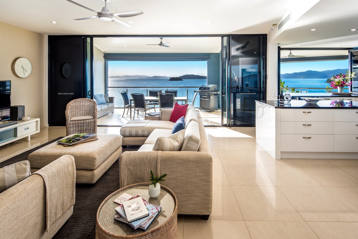 A spacious open-plan living area features modern furnishings in neutral tones. Floor-to-ceiling glass doors provide stunning views of the water and distant islands. A comfortable seating arrangement includes a large sectional sofa and armchairs, while a dining table is set for outdoor meals on the adjoining balcony.