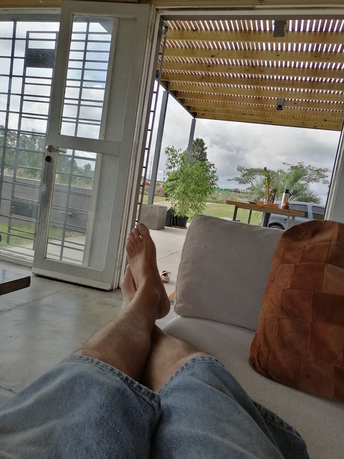 A comfortable seating area is visible, featuring a light beige sofa and natural light filtering through large doors. A view of a grassy outdoor space can be seen in the background, complemented by greenery and a shaded patio area, creating an inviting atmosphere.