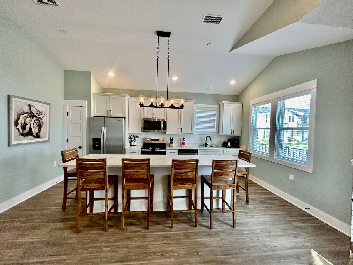 The kitchen area features a large island with seating for six, complemented by modern light fixtures above. White cabinetry stands against a soft green wall, while stainless steel appliances are neatly arranged. Natural light enters from windows, enhancing the inviting atmosphere.