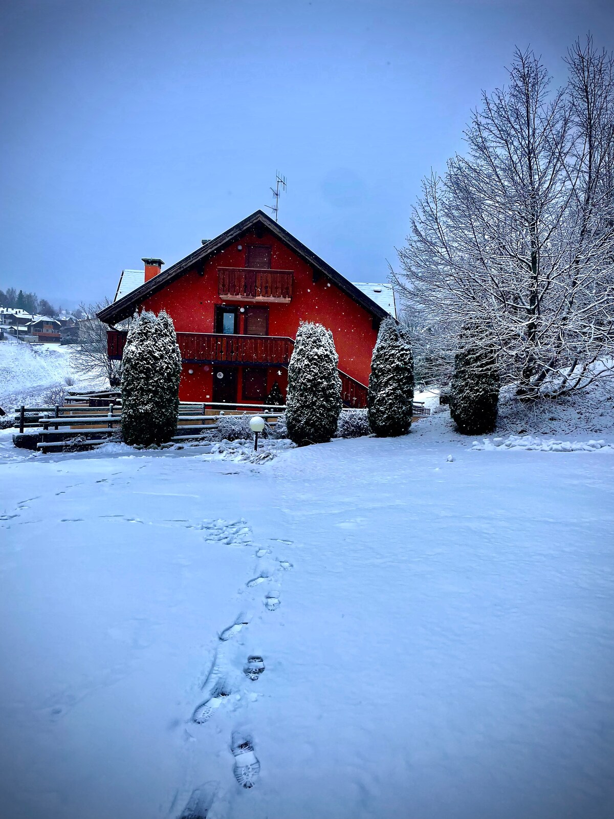 A two-story red house is nestled in a snowy landscape, surrounded by tall, green conifers. A light dusting of snow covers the ground, with fresh footprints leading towards the house. Soft gray skies create a serene winter atmosphere.