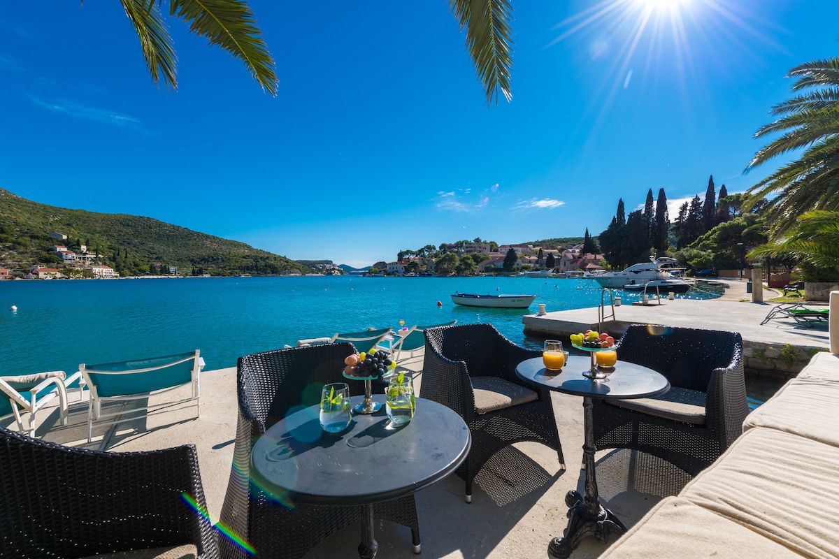 A seating area is presented by the water's edge, featuring dark wicker chairs and round tables. Refreshments on the tables provide a relaxed setting. The tranquil sea reflects the clear blue sky, with boats anchored in the distance and palm trees enhancing the coastal view.