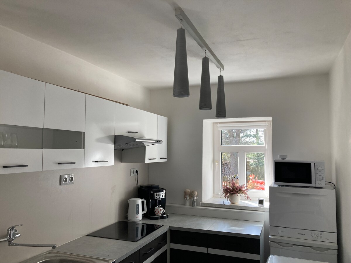 A modern kitchen features sleek cabinetry in a white finish, complemented by a grey countertop. Three pendant lights hang from the ceiling above the work area, illuminating the cooking space. A window with a view allows natural light to fill the room.