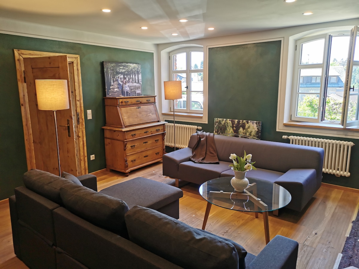 A spacious living room features two gray sofas facing a glass coffee table. Natural light enters through large windows, highlighting the wooden accents, including a vintage writing desk. A modern lamp provides additional soft light, complemented by a green wall and decorative plant.