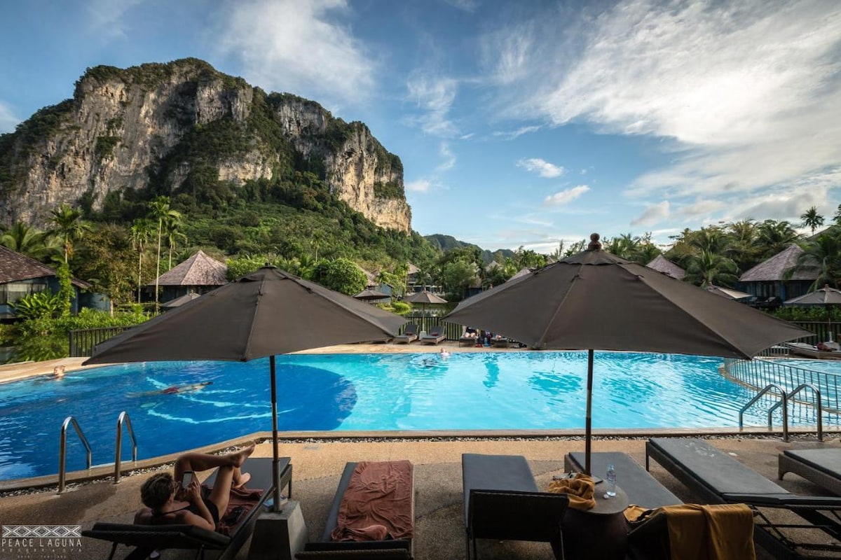 The outdoor swimming pool is surrounded by sun loungers and large umbrellas, providing shaded areas for relaxation. Lush greenery and towering cliffs are visible in the background, creating a serene environment. The clear blue water glistens under a bright sky, inviting guests to unwind.