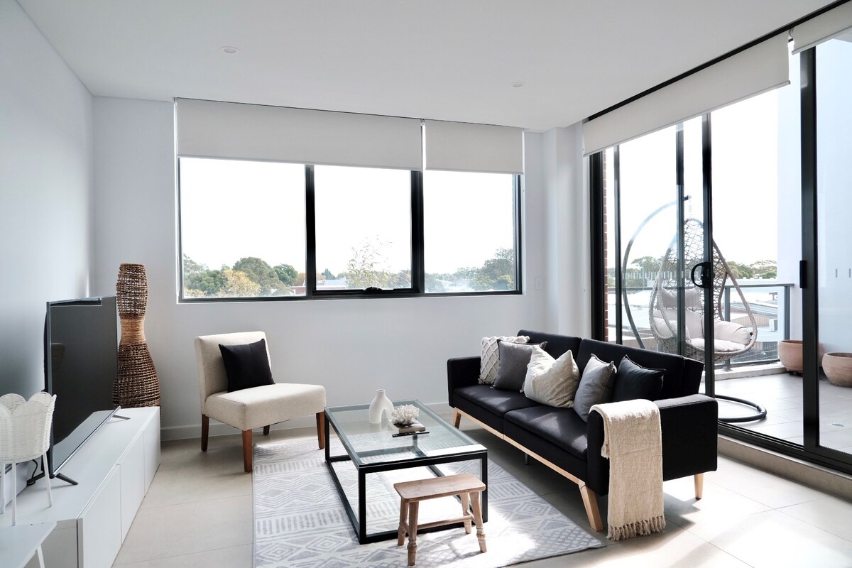 A modern living space showcases large windows that provide natural light and views of the surroundings. The area features a comfortable couch, an accent chair, and a glass coffee table. A stylish decorative piece stands nearby, while a balcony is visible through the sliding door.