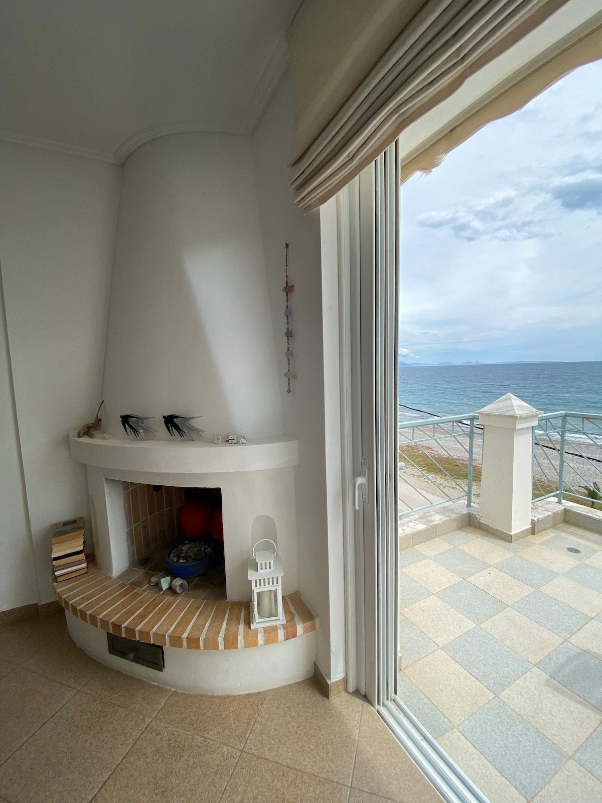 A cozy fireplace is positioned in the corner of a bright interior space, complementing the large glass door that opens onto a balcony with sea views. The brick base of the fireplace adds a subtle touch of warmth to the room, contrasting with the cool tones of the tiles and ocean.