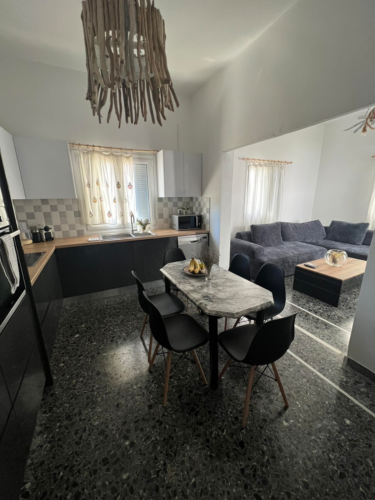 A modern kitchen and dining area feature sleek cabinetry and a marble-topped table surrounded by black chairs. Natural light filters through the window, illuminating the space. A comfortable living room is visible in the background, furnished with dark sofas and decorative accents.